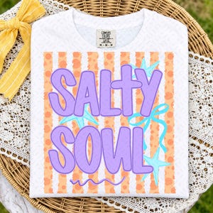 May include: A white short-sleeved t-shirt featuring a graphic print with the text SALTY SOUL in large purple letters. The background of the graphic consists of vertical orange stripes with polka dots, accented by light blue starfish and a ribbon.
