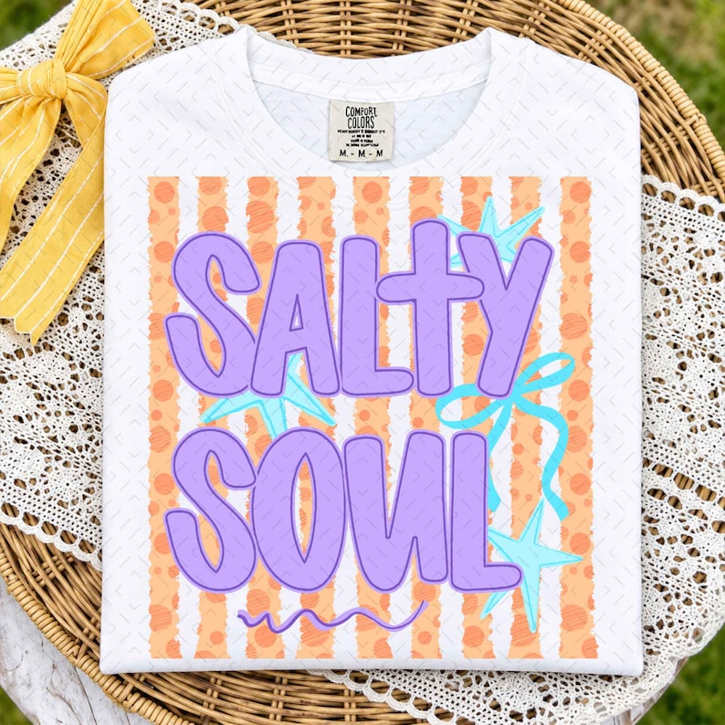 May include: A white short-sleeved t-shirt featuring a graphic print with the text SALTY SOUL in large purple letters. The background of the graphic consists of vertical orange stripes with polka dots, accented by light blue starfish and a ribbon.
