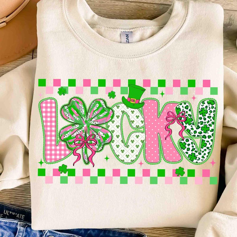 May include: Cream-colored sweatshirt with the word "LUCKY" in a playful, St. Patrick's Day-themed design. The letters are in pink and green, with a clover and a leprechaun hat. The design includes pink and green checkered patterns.