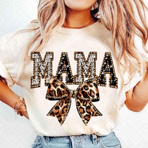 May include: Cream-colored t-shirt with the word "MAMA" in jeweled letters. The "M" and "A" are black with crystal accents, while the second "A" is silver with crystal accents. A leopard print bow is centered below the word.