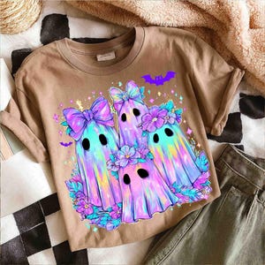 Op de afbeelding: Een bruin T-shirt met een kleurrijke afbeelding van drie spookkarakters met strikken, bloemen en vleermuisaccenten. De geesten hebben een regenbooggradi&euml;ntontwerp en zwarte ogen. Het ontwerp is omgeven door bloemen- en sterrendetails.