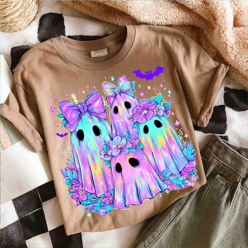 Op de afbeelding: Een bruin T-shirt met een kleurrijke afbeelding van drie spookkarakters met strikken, bloemen en vleermuisaccenten. De geesten hebben een regenbooggradi&euml;ntontwerp en zwarte ogen. Het ontwerp is omgeven door bloemen- en sterrendetails.