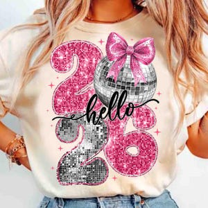 May include: A cream-colored t-shirt featuring a design with the numbers "2026" in pink glitter, a disco ball, and a pink bow. The word "hello" is written in cursive script. The design includes small pink star accents.