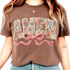 May include: A brown t-shirt with the word "AMEN" embroidered in a floral design. The letters are filled with pink, yellow, and blue flowers. A pink knitted bow is below the word.