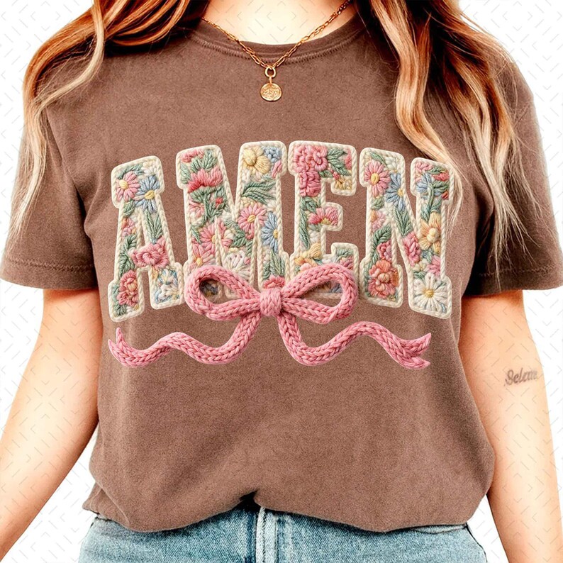 May include: A brown t-shirt with the word "AMEN" embroidered in a floral design. The letters are filled with pink, yellow, and blue flowers. A pink knitted bow is below the word.