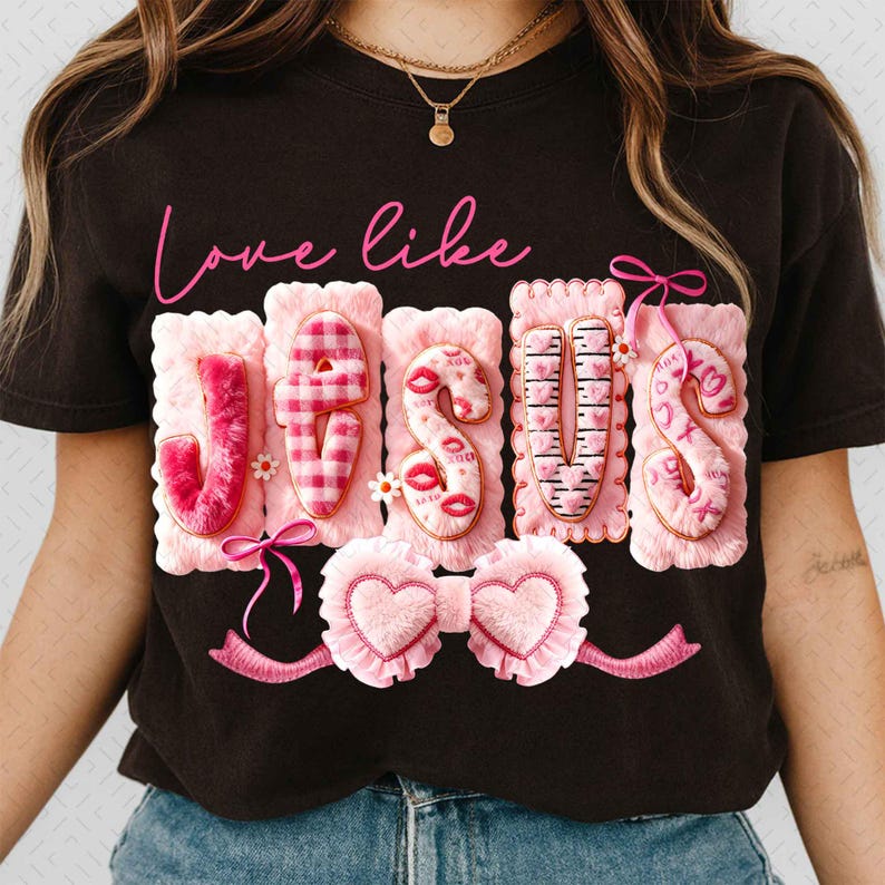 Op de afbeelding: Zwart T-shirt met de roze tekst "Love like Jesus". Het woord "JESUS" is weergegeven in getextureerde roze letters met decoratieve elementen zoals lippen, harten en ruitpatronen. Onderaan een roze hartvormige strik.