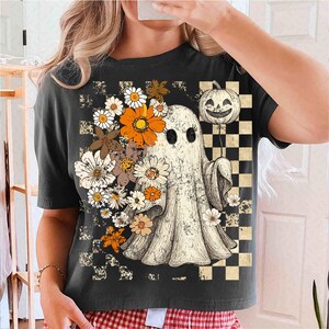 May include: Dark gray t-shirt featuring a vintage-style ghost with a pumpkin balloon and floral accents. The ghost is surrounded by daisies and orange flowers. A checkered pattern is on the right side of the shirt. The shirt has a crew neck and short sleeves.