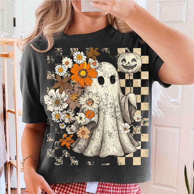 May include: Dark gray t-shirt featuring a vintage-style ghost with a pumpkin balloon and floral accents. The ghost is surrounded by daisies and orange flowers. A checkered pattern is on the right side of the shirt. The shirt has a crew neck and short sleeves.