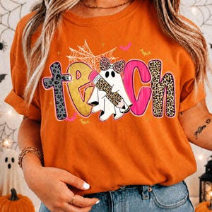 Peut inclure: Un t-shirt orange avec le mot TEACH &eacute;crit en grandes lettres color&eacute;es. Le T est en imprim&eacute; l&eacute;opard gris, le E est dor&eacute;, le A est un fant&ocirc;me blanc tenant un crayon l&eacute;opard, le C est rose et le H est en imprim&eacute; l&eacute;opard. Une toile d'araign&eacute;e est au-dessus.