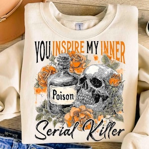May include: Cream-colored sweatshirt with a graphic design featuring a skull, a poison bottle, and orange roses. The text reads "You Inspire My Inner Serial Killer" in black and orange lettering. The design is centered on the front of the sweatshirt.