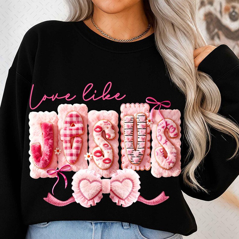 Op de afbeelding: Zwart sweatshirt met de tekst "Love like Jesus" in roze script. Het woord "Jesus" is geschreven met roze, getextureerde letters, elk versierd met unieke ontwerpen, waaronder lippen, harten en een liniaal. Een roze strik en lint maken het ontwerp compleet.