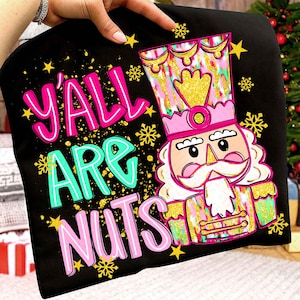May include: Black t-shirt with the text "Y'all Are Nuts" in pink and teal. A colourful nutcracker graphic is featured on the right, with gold, pink, and green accents. Yellow stars and snowflakes complete the festive design.