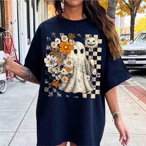 May include: Navy blue t-shirt featuring a vintage-style ghost design with flowers and a pumpkin. The graphic includes a checkered pattern and a distressed effect, creating a retro Halloween aesthetic.