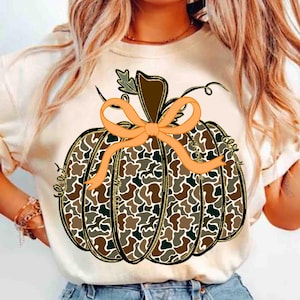May include: A cream-colored t-shirt featuring a large pumpkin design. The pumpkin has a camouflage pattern in shades of brown, green, and tan, outlined in black and gold. An orange bow adorns the top, with a brown stem and green leaf. The design is centered on the shirt.