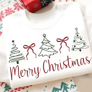 May include: White sweatshirt with a green Christmas tree design and red bows. The words "Merry Christmas" are written in red script below the trees. A festive holiday-themed top.
