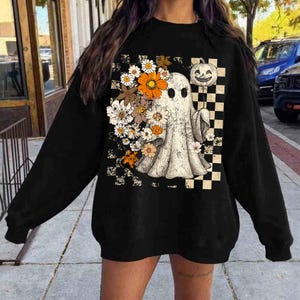 May include: Black sweatshirt featuring a vintage-style ghost design holding a pumpkin and flowers. The ghost is surrounded by a checkered pattern. The design includes orange, white, and beige colors.