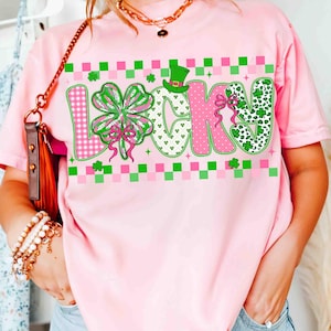 May include: Pink t-shirt with the word "LUCKY" in a colorful design. The "L" features a pink and white checkered pattern, the "U" has a four-leaf clover, the "C" is pink, and the "K" and "Y" have green and white patterns. A green hat is above the "C".