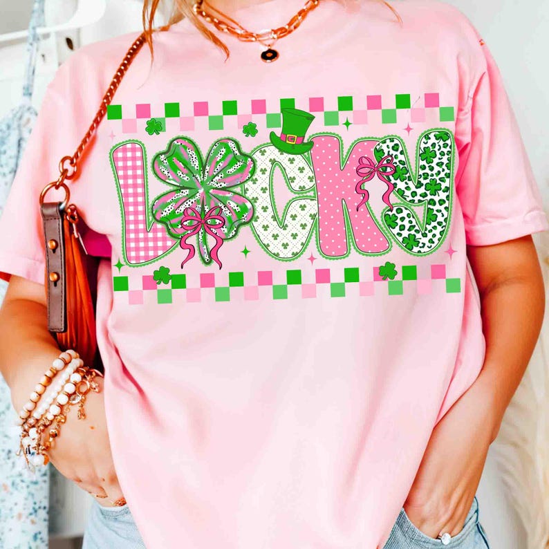 May include: Pink t-shirt with the word "LUCKY" in a colorful design. The "L" features a pink and white checkered pattern, the "U" has a four-leaf clover, the "C" is pink, and the "K" and "Y" have green and white patterns. A green hat is above the "C".