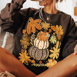 May include: A dark brown sweatshirt featuring a fall-themed graphic. The design includes stacked pumpkins with a leopard print base, orange and yellow flowers, and a small bow. The sweatshirt is a long-sleeved, crew neck style.
