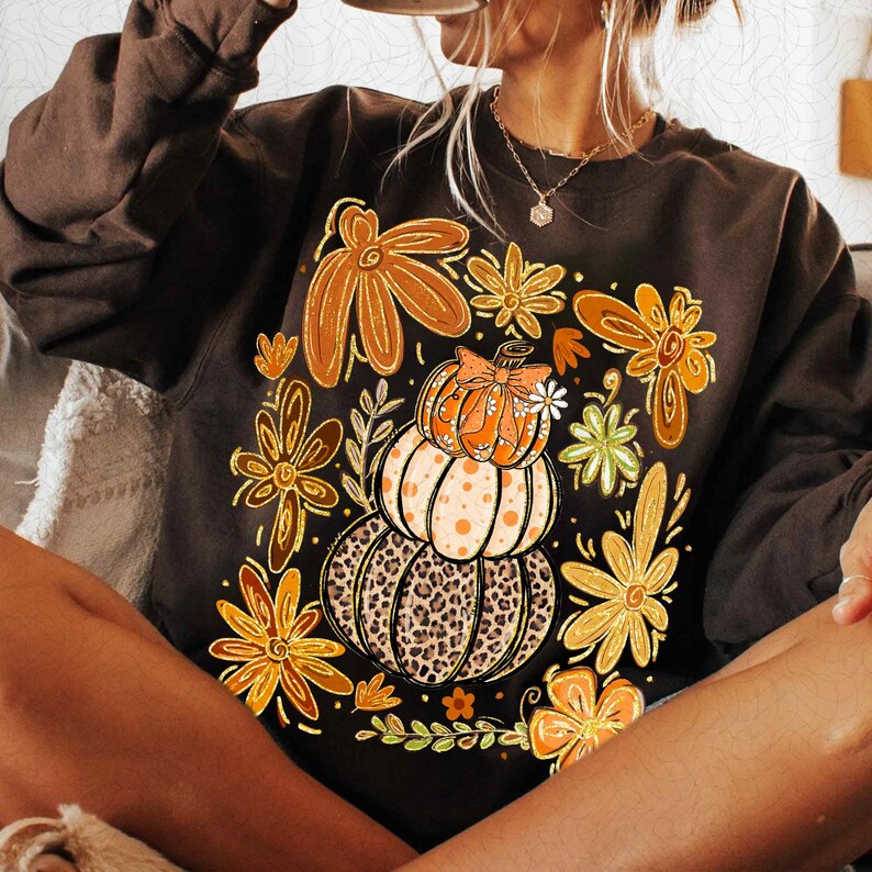 May include: A dark brown sweatshirt featuring a fall-themed graphic. The design includes stacked pumpkins with a leopard print base, orange and yellow flowers, and a small bow. The sweatshirt is a long-sleeved, crew neck style.