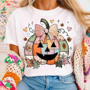 May include: White t-shirt featuring a patchwork pumpkin design with a smiling face, bow, and floral accents. The pumpkin has a green stem and is surrounded by hearts and leaves, perfect for the fall season.
