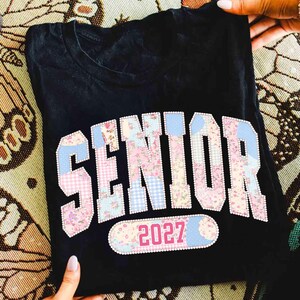 Peut inclure: T-shirt noir avec le mot "SENIOR" dans un motif patchwork floral rose, vichy bleu et accents blancs. L'ann&eacute;e "2027" est affich&eacute;e en dessous. Le t-shirt est pli&eacute;.