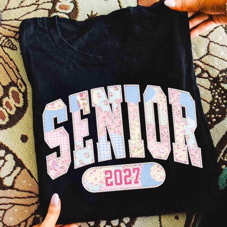 Peut inclure: T-shirt noir avec le mot "SENIOR" dans un motif patchwork floral rose, vichy bleu et accents blancs. L'ann&eacute;e "2027" est affich&eacute;e en dessous. Le t-shirt est pli&eacute;.