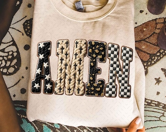 May include: Cream-colored sweatshirt with the word "AMEN" in a patchwork design. The letters feature various patterns: stars, crosses, leopard print, and a checkered pattern. The sweatshirt is held by hands.