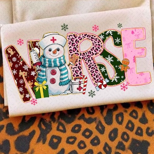 Op de afbeelding: Een crèmekleurig t-shirt met het woord "NURSE" in feestelijke letters. De "N" is versierd met peperkoekmannetjes en sneeuwvlokken, de "R" met een luipaardprint, de "S" met sneeuwvlokken en de "E" met roze strikken. In het midden staat een sneeuwman met een verpleegstersmuts en sjaal.