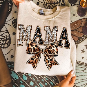 May include: A beige sweatshirt with the word "MAMA" in jeweled letters and a leopard print bow. The letters are outlined with silver and black gems. The bow is brown and black with a leopard print pattern.