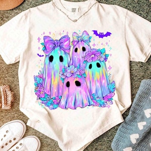 Op de afbeelding: Wit t-shirt met een kleurrijke illustratie van vier spookkarakters met strikken en bloemenkransen. De geesten hebben een regenboogkleurenschema met paarse, blauwe en roze tinten. Een paarse vleermuisafbeelding staat erboven.