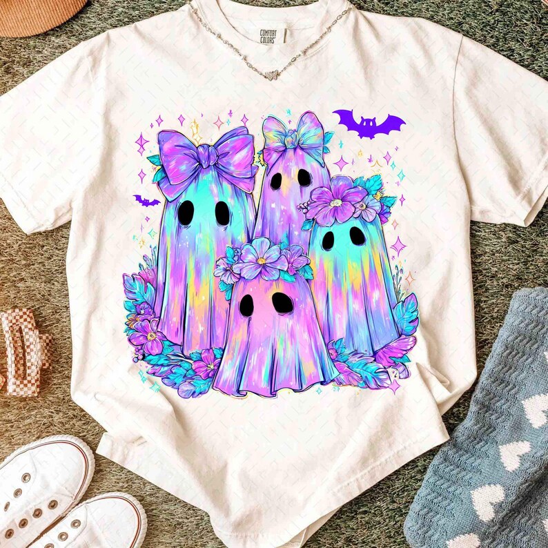 Op de afbeelding: Wit t-shirt met een kleurrijke illustratie van vier spookkarakters met strikken en bloemenkransen. De geesten hebben een regenboogkleurenschema met paarse, blauwe en roze tinten. Een paarse vleermuisafbeelding staat erboven.