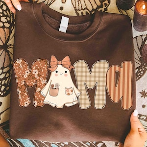 May include: A brown sweatshirt with the word "MAMA" in a patchwork design. The "M" is floral, the "A"s are plaid and striped, and the ghost-shaped "A" has a bow and flower pocket.