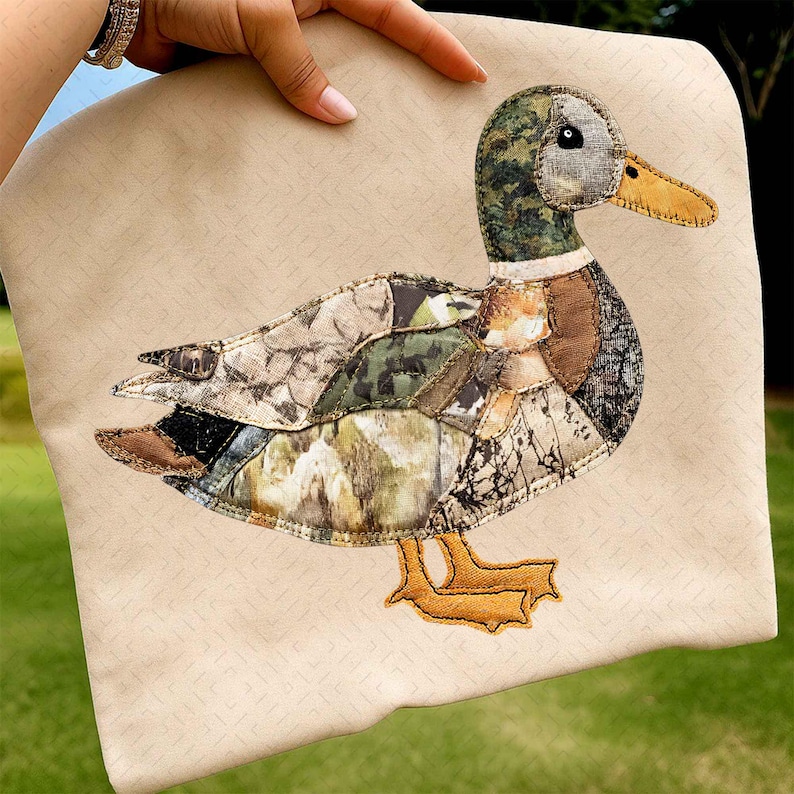 May include: A beige pillow with a detailed appliqu&eacute; of a duck. The duck features a patchwork design with camouflage and brown fabrics, a yellow beak, and orange feet. The pillow is held by a hand.