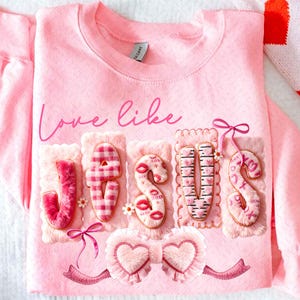Op de afbeelding: Zachtroze sweatshirt met de tekst "Love like Jesus". Het woord "Jesus" is geschreven met pluche, getextureerde letters, elk met unieke patronen en versieringen, waaronder harten en lippen. Strikken en hartaccenten maken het ontwerp compleet.