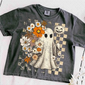 May include: Dark grey t-shirt featuring a vintage-style illustration of a ghost adorned with flowers and holding a jack-o'-lantern balloon. The design includes a checkered background and a retro aesthetic, perfect for Halloween.