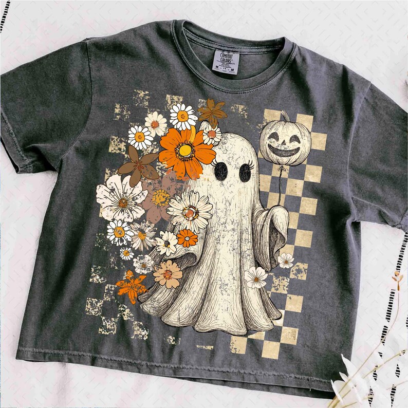 May include: Dark grey t-shirt featuring a vintage-style illustration of a ghost adorned with flowers and holding a jack-o'-lantern balloon. The design includes a checkered background and a retro aesthetic, perfect for Halloween.