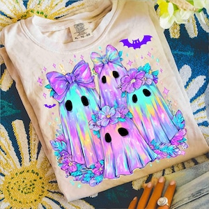Op de afbeelding: Een beige sweatshirt met een kleurrijke afbeelding van drie spookfiguren met strikken en bloemenkransen. De geesten hebben zwarte ovale ogen en zijn omringd door paarse bloemen en vleermuizen. Het shirt heeft een Comfort Colors-label.