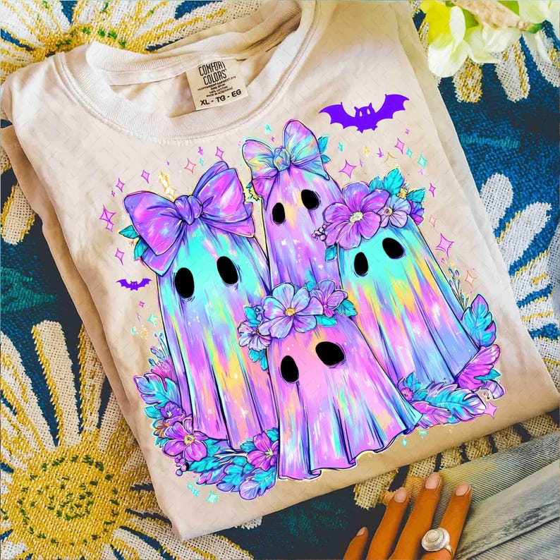 Op de afbeelding: Een beige sweatshirt met een kleurrijke afbeelding van drie spookfiguren met strikken en bloemenkransen. De geesten hebben zwarte ovale ogen en zijn omringd door paarse bloemen en vleermuizen. Het shirt heeft een Comfort Colors-label.