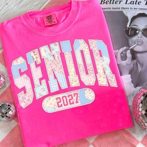 Peut inclure: Un sweat-shirt rose vif avec le mot "SENIOR" dans un motif patchwork de motifs floraux et vichy. L'ann&eacute;e "2027" est en dessous. Le sweat-shirt est de la marque Comfort Colors.