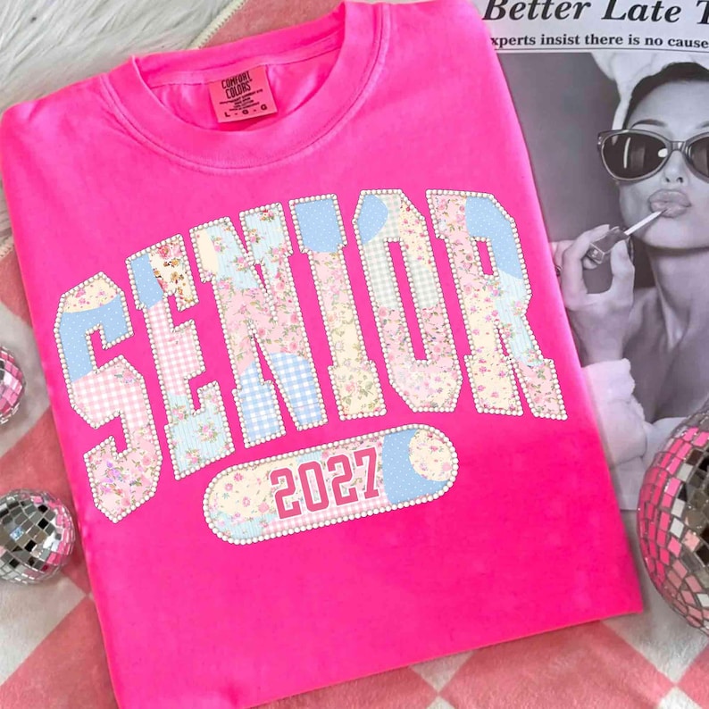 Peut inclure: Un sweat-shirt rose vif avec le mot "SENIOR" dans un motif patchwork de motifs floraux et vichy. L'ann&eacute;e "2027" est en dessous. Le sweat-shirt est de la marque Comfort Colors.