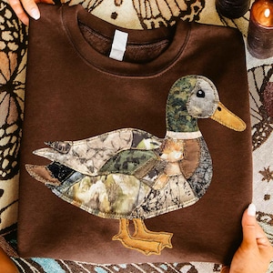 May include: A brown sweatshirt featuring an appliqued duck design. The duck is made of various camouflage fabric patches, with a green and brown head, a yellow beak, and yellow feet. The sweatshirt is a solid brown color.
