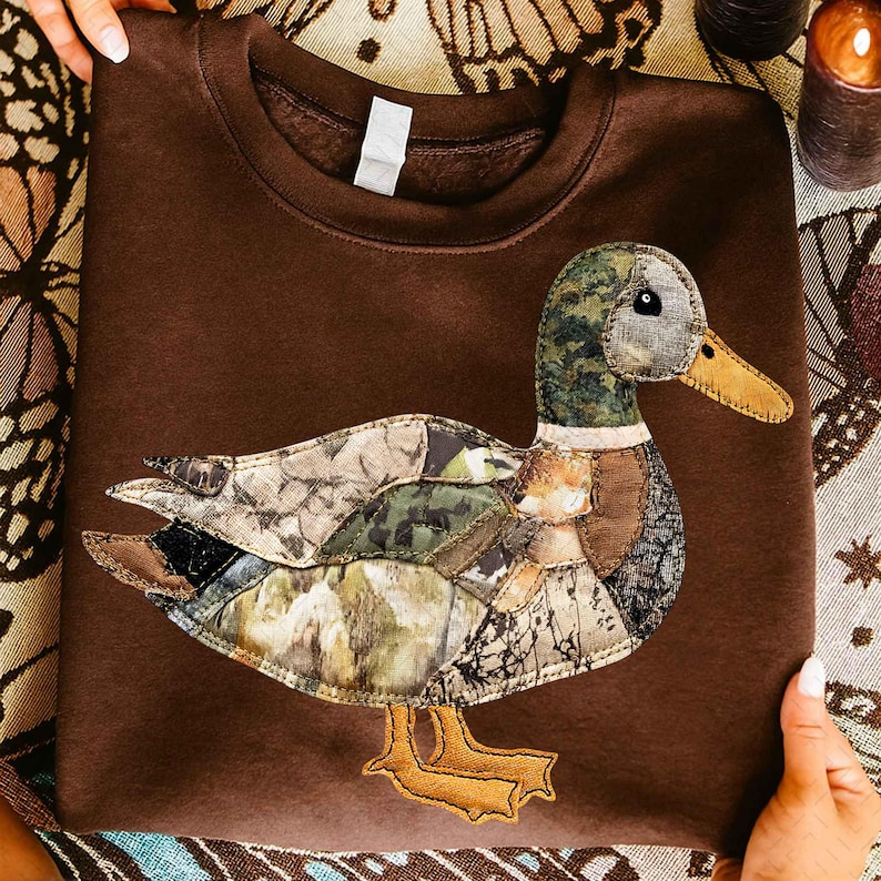 May include: A brown sweatshirt featuring an appliqued duck design. The duck is made of various camouflage fabric patches, with a green and brown head, a yellow beak, and yellow feet. The sweatshirt is a solid brown color.