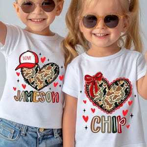 May include: Two children wearing white t-shirts with heart designs. One shirt has a heart with a camouflage pattern, a baseball cap, and the name "Jameson". The other shirt has a heart with a camouflage pattern, a red bow, and the name "Chip".
