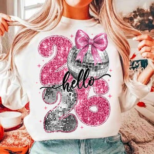 May include: White sweatshirt with a graphic design featuring the numbers "26" in pink glitter, a disco ball, and a pink bow. The word "hello" is written in black script across the design. Pink stars are scattered around the design.
