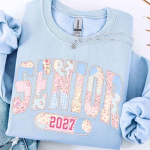 Peut inclure: Un sweat-shirt bleu clair avec le mot "SENIOR" dans un motif patchwork avec des motifs floraux et vichy. L'ann&eacute;e "2027" est en dessous du mot "SENIOR". Un collier argent&eacute; avec un pendentif fleur est sur le sweat-shirt.