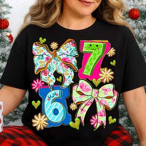 May include: Black t-shirt featuring the numbers 6 and 7, along with two floral bows and two additional bows with floral patterns. The design includes hearts and flowers in various colors, creating a festive and celebratory theme.