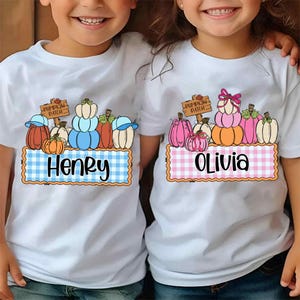 May include: White t-shirts featuring a colorful pumpkin patch design. One shirt has a blue and white checkered name tag that says "Henry". The other shirt has a pink and white checkered name tag that says "Olivia".