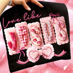 Op de afbeelding: Zwarte stof met de woorden "Love like Jesus" in roze script. Het woord "Jesus" is gespeld in roze, pluizige letters met decoratieve elementen zoals lippen, harten en ruitpatronen. Een roze strik en hartvormige accenten staan onderaan.