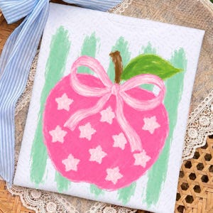 Peut inclure: Un carré de tissu blanc orné d'une pomme rose peinte avec une tige brune et une feuille verte. La pomme est décorée d'un nœud rose clair et de plusieurs étoiles rose clair. Le fond présente des rayures verticales en coups de pinceau vert clair.